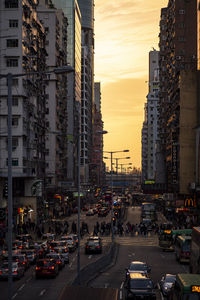 Traffic on city street and buildings against sky at sunset