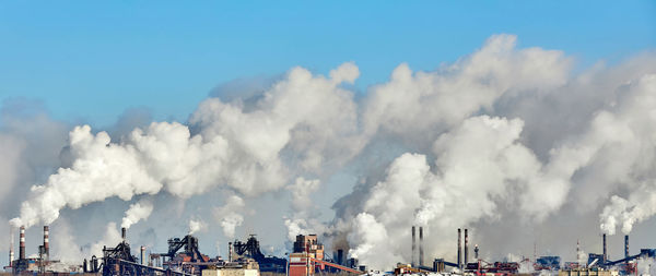 Panoramic view of smoke emitting from factory against blue sky