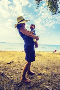 Woman carrying baby while standing on beach against sea