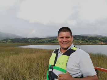 Portrait of smiling man standing on field by lake against sky
