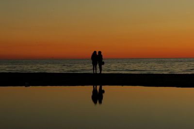 Silhouette people standing on beach against orange sky
