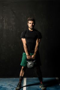 Full length powerful male athlete lifting heavy kettlebell while standing against black wall during functional workout in gym