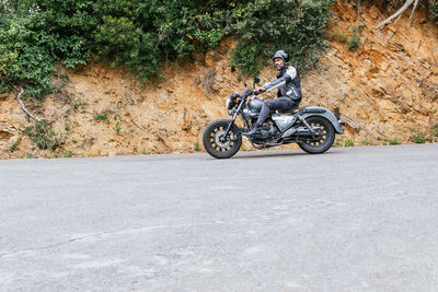 Full body of focused bearded ethnic male biker in black leather jacket and helmet riding modern motorbike on asphalt road amidst lush green trees growing in mountainous valley