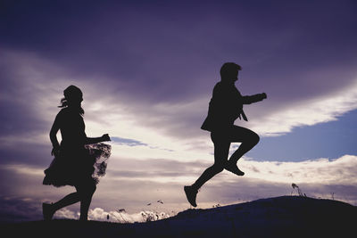 Silhouette man with woman jumping against cloudy sky during sunset