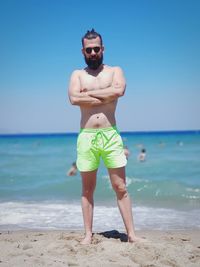 Portrait of shirtless man standing at beach against blue sky