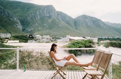 Woman sitting on chair by lake against mountains