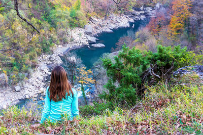 A young girl with long hair in a light turquoise blouse sits on a cliff and looks at the river