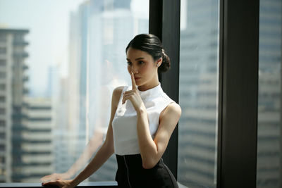 Businesswoman with finger on lips standing against window at office