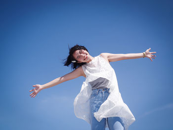 Low angle portrait of happy young woman with arms outstretched standing against blue sky