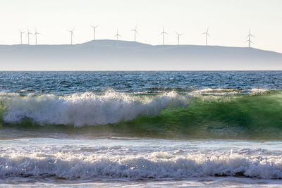Waves rushing towards shore with wind turbines on mountain in background