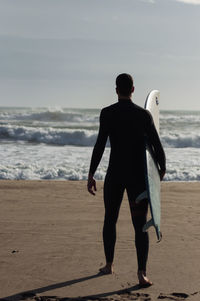 Rear view of man holding surfboard while standing at beach