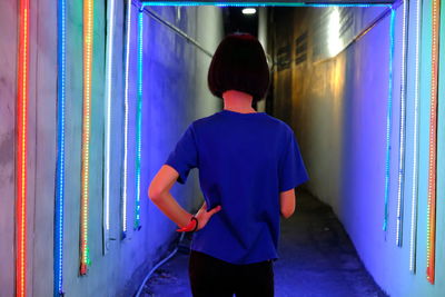 Rear view of young woman standing by illuminated wall