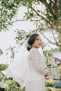 Bride looking away while standing by tree