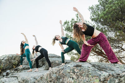 Female friends practicing yoga on rocks against clear sky during weekend