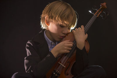 Boy holding violin while sitting against black background