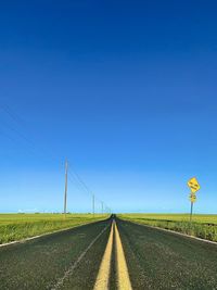 Country road amidst field against clear blue sky