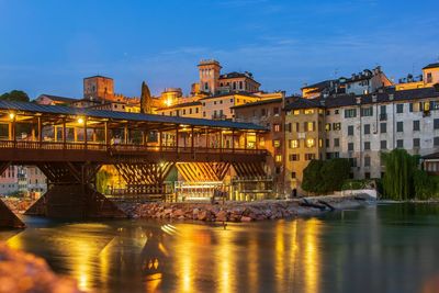 Illuminated bridge over river by buildings against sky at night