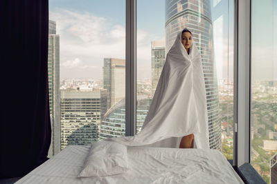 Young woman wrapped in a sheet stands in a bedroom in a skyscraper during the day