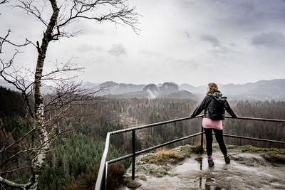 Rear view of woman standing on railing with mountains in background in fog and rain