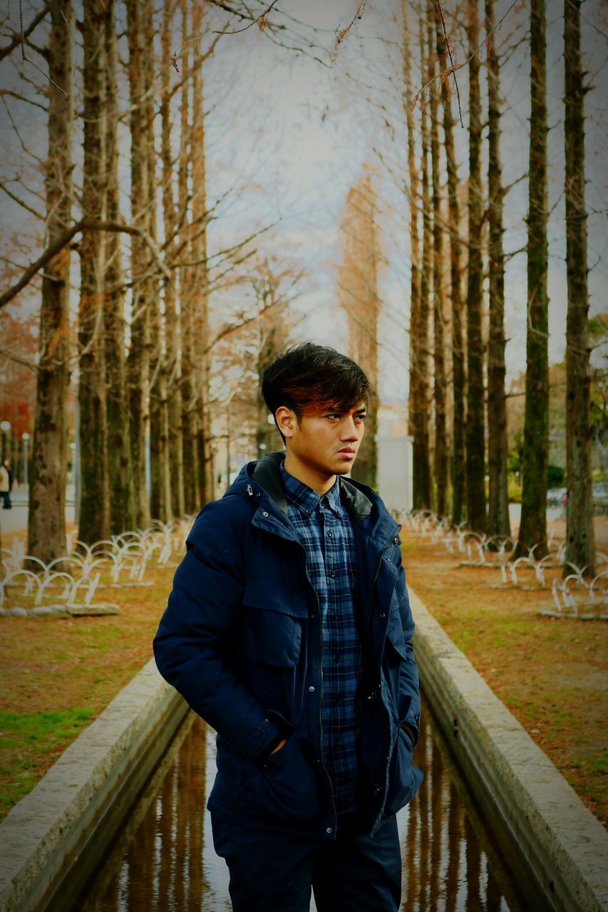 tree, one person, standing, young men, young adult, plant, real people, forest, land, lifestyles, three quarter length, leisure activity, trunk, tree trunk, front view, day, clothing, casual clothing, nature, woodland, outdoors, teenager, warm clothing, teenage boys, adolescence
