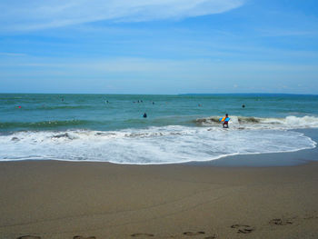 Mid distance view of man holding surfboard on shore at beach against blue sky