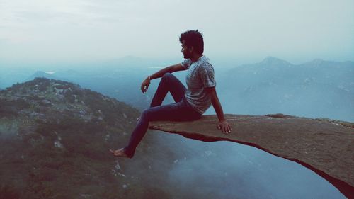 Full length of man sitting on cliff against mountain during foggy weather