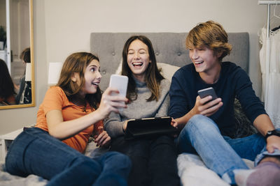 Smiling friends using technology while sitting on bed at home