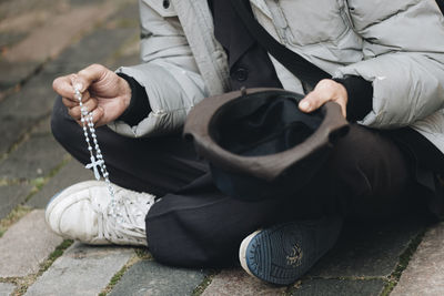 Low section of man holding hat and crucifix while sitting on footpath