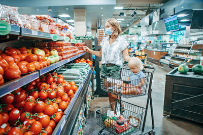 Young mother in protective face mask buying food with kid baby in shopping cart. 