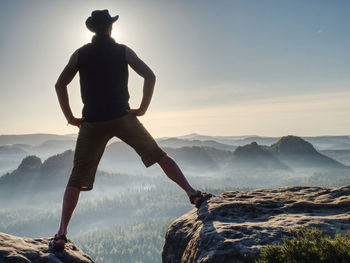 Cowboy style hiker man on cliff watching over misty morning valley to sun. man body with akimbo arms