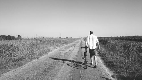 Full length rear view of man with bicycle walking on road against sky during sunny day