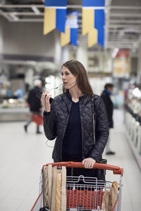 Woman talking on mobile phone through headphones while standing with shopping cart at supermarket