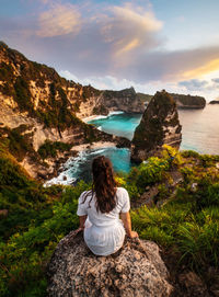 Rear view of woman sitting on rock by sea against sky