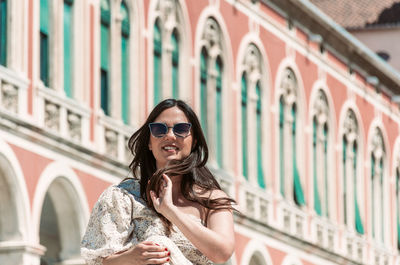 Portrait of beautiful young woman with long dark hair, sitting in square with beautiful architecture