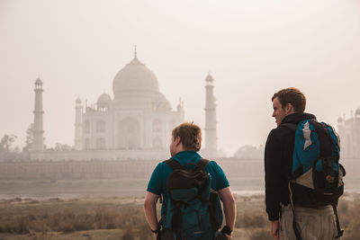 Rear view of tourists carrying backpacks while standing in front of taj mahal during foggy weather