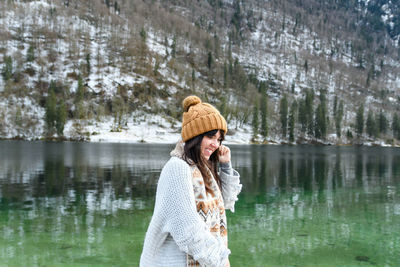 Young woman in winter sweater smiling, standing by a lake.