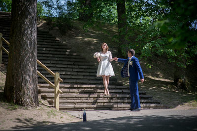 Full length of wedding couple holding hands on steps at park during sunny day