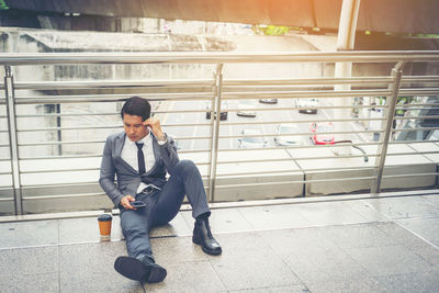 Thoughtful businessman using phone while sitting on elevated walkway in city 