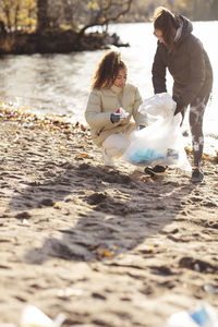 Young female environmentalist with teenage girl collecting waste while crouching on land