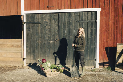 Full length portrait of female farmer with shovel standing by organic vegetables outside barn