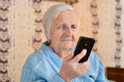 Happy elderly 90-year-old woman with glasses wearing a blue jacket smiles using a smartphone.