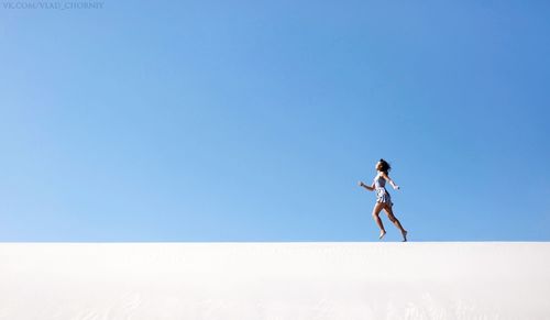 Side view of mid adult woman running on sand at desert against clear blue sky