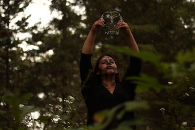 Low angle view of young woman holding mask while standing amidst plants