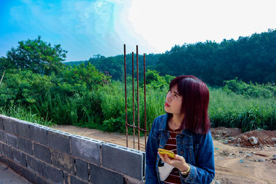 Thoughtful woman with mobile phone sitting by brick wall against trees