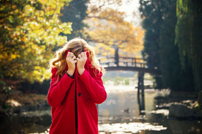 Young woman hiding under red coat in autumn city park near lake