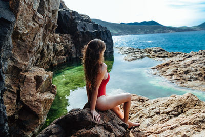 Side view of seductive woman wearing swimsuit while sitting on rock by sea