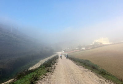 Rear view of men cycling on dirt road against sky during foggy weather