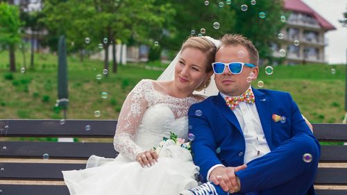 Bride and groom looking away while sitting on bench at park