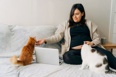 A middle-aged pregnant woman sits on the couch at home and pets cats.