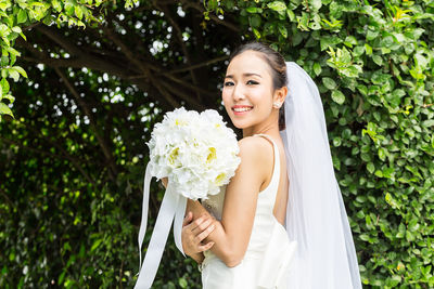 Portrait of smiling bride with bouquet standing against plants 